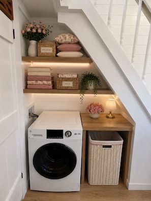 A laundry area with a washing machine, a drying rack, and storage shelves with folded towels and baskets.