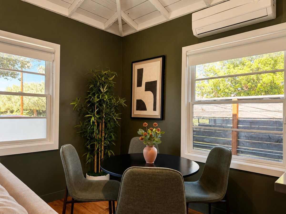 A room with a dining table, four chairs, a potted plant, and a framed artwork on the wall.