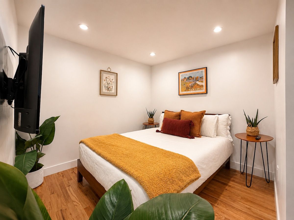 A bedroom with a bed, a television, two wall-mounted paintings, a small side table, and a potted plant.