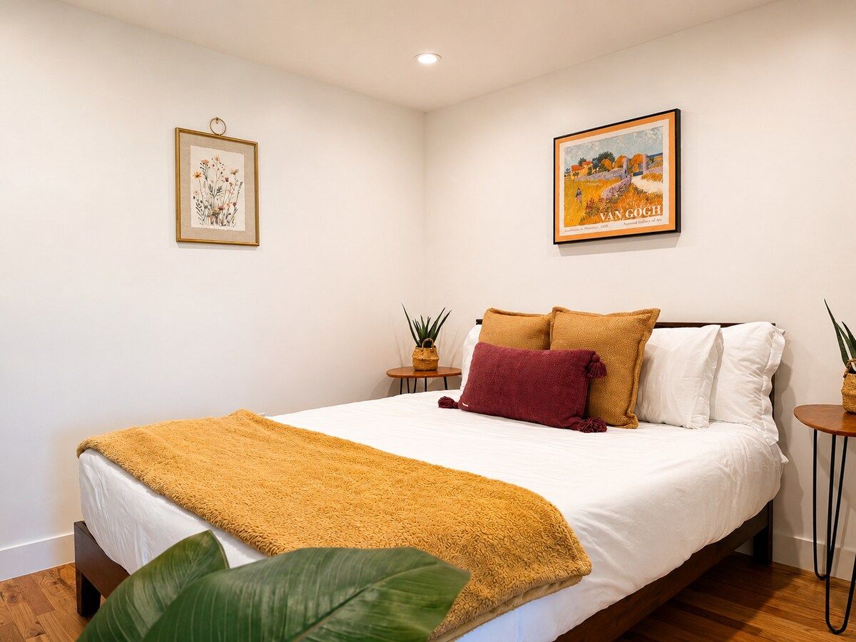 A bedroom with a bed, pillows, a blanket, two bedside tables with plants, and two framed pictures on the walls.