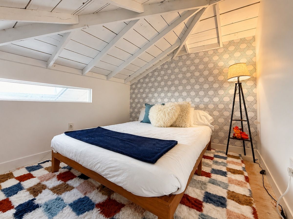 A bedroom with a sloped ceiling, a bed with white bedding, a patterned rug, and a floor lamp.