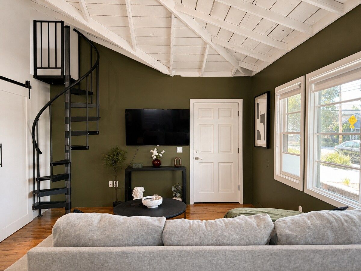 A modern living room with a staircase, a flat-screen TV, a sofa, and a window with a view.
