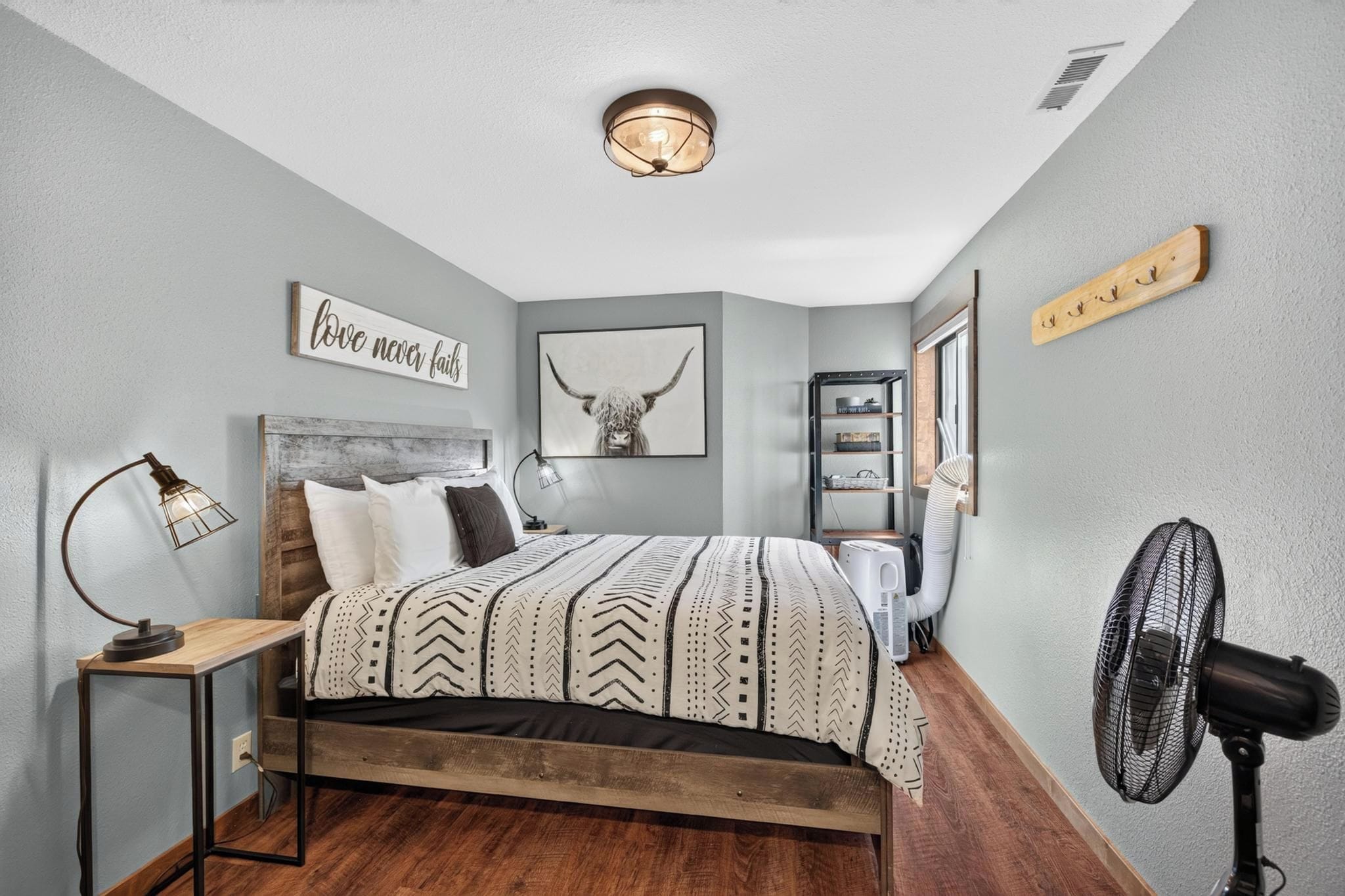 A bedroom with a bed, bedside table, lamp, wall art, and a standing fan.