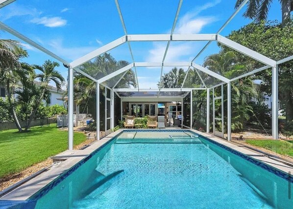A swimming pool with a covered area, surrounded by greenery and a clear sky.