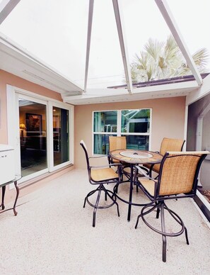A covered patio area with a round table and chairs, a glass door leading to an indoor space, and a view of palm trees outside.