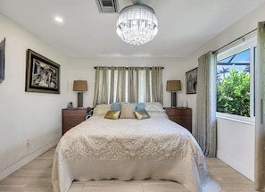 A bedroom with a large bed, two bedside tables, a chandelier, and a window with curtains.