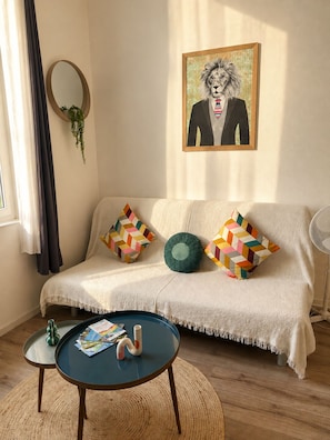 Un salon chaleureux avec un canapé, des coussins géométriques aux couleurs vives, une table basse ronde sur laquelle sont posés des magazines, une plante suspendue et une œuvre d’art encadrée accrochée au mur.