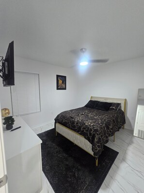 Een slaapkamer met een bed, een bureau, een plafondventilator en een aan de muur bevestigde televisie.