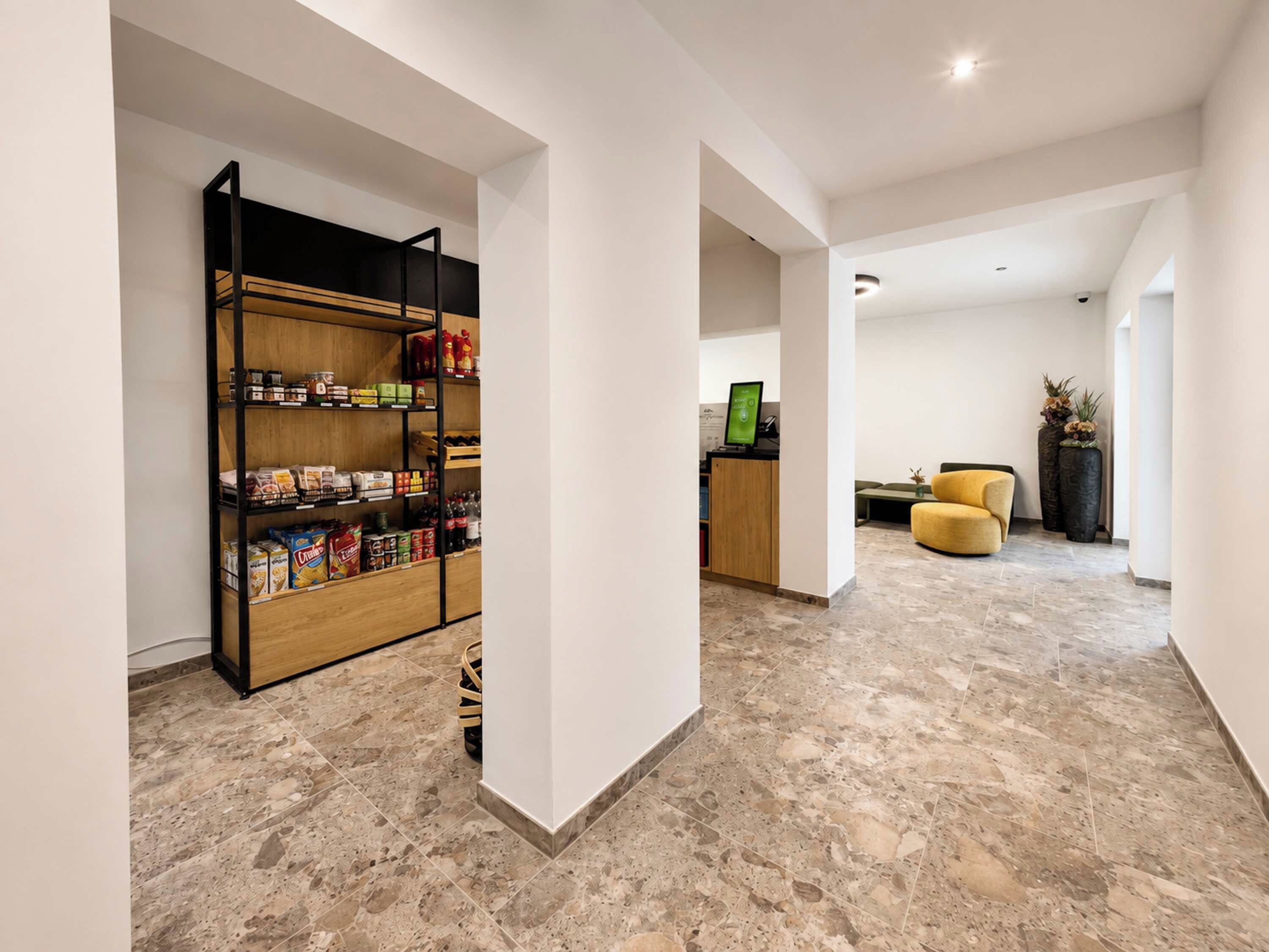 Ein modernes Interieur mit Küchenzeile, an der sich ein Vorratsschrank aus Holz mit Lebensmitteln, eine Sitzecke mit einem gelben Sessel und ein Empfangsschalter mit Computerbildschirm befinden.
