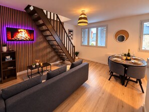 A modern living room with a fireplace, a staircase, a dining area with a round table, and a large window. - The Duplex des Prébendes - Central (Tours)
