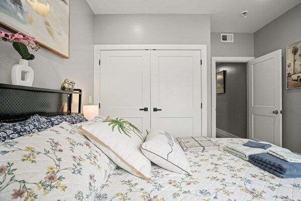 A bedroom with a bed, a nightstand, a wall-mounted shelf with an owl figurine, a framed painting, and a door leading to another room.