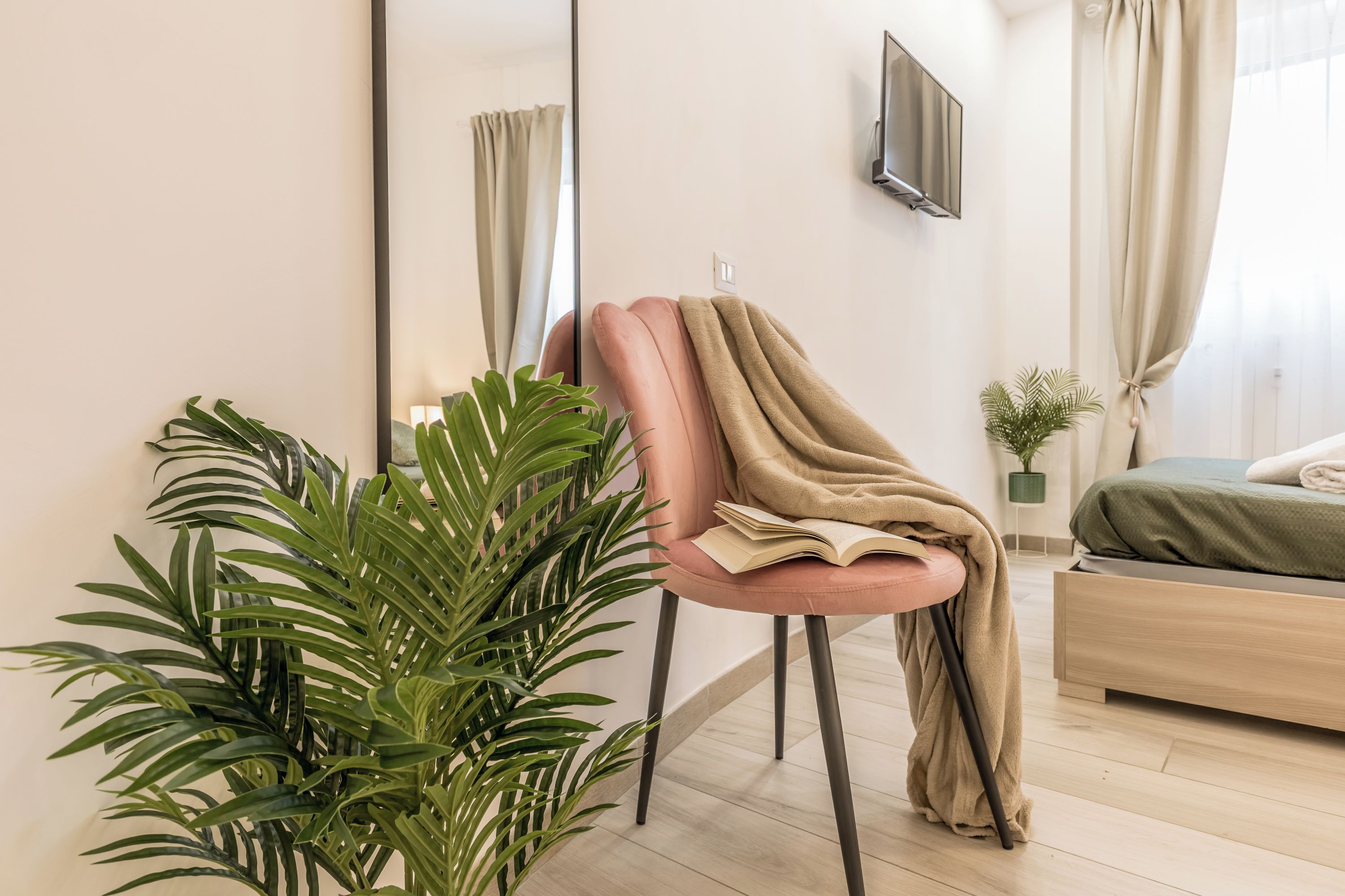 Un salon moderne avec un fauteuil rose, un grand miroir, un téléviseur à écran plat, une plante verte et un lit recouvert d’une couverture verte.