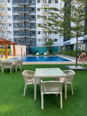 Piscina com mesa de vidro e cadeiras de vime, rodeada por grama sintética, em frente a um edifício com vários andares.