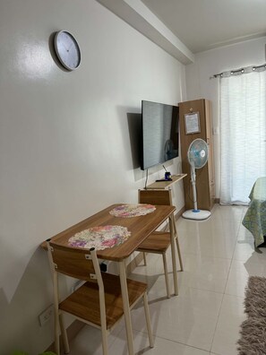 Uma sala de jantar com mesa de madeira, cadeiras, relógio fixado na parede, televisão e ventilador.