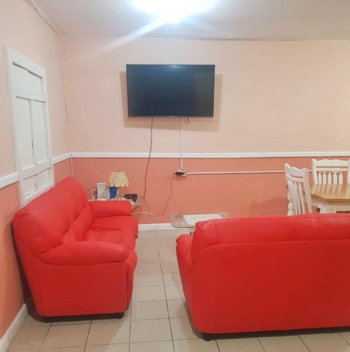 Et værelse med to røde sofaer, et tv ophængt på væggen, et lille bord med en lampe og en hvid stol.