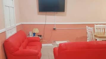 Et værelse med to røde sofaer, et tv ophængt på væggen, et lille bord med en lampe og en hvid stol.