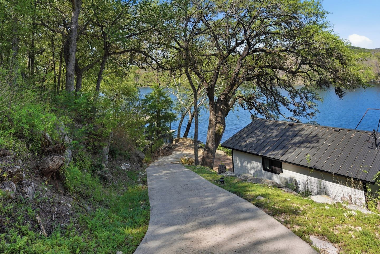 A pathway leading to a small building with a metal roof, surrounded by trees and a body of water.