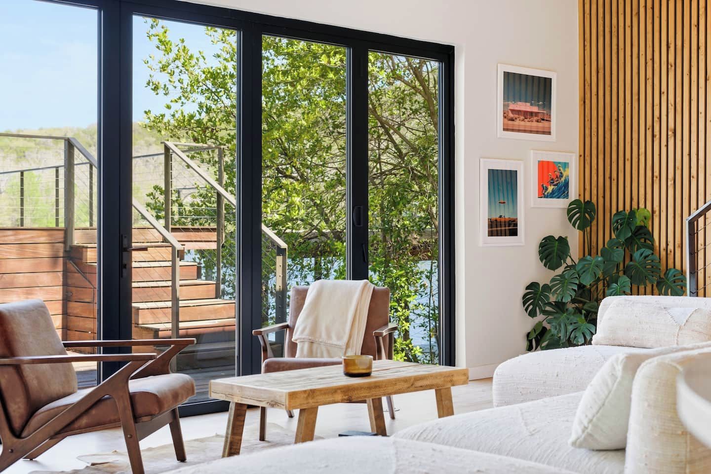 A modern living room with a view of a wooden deck and greenery, a staircase, and artwork on the wall.