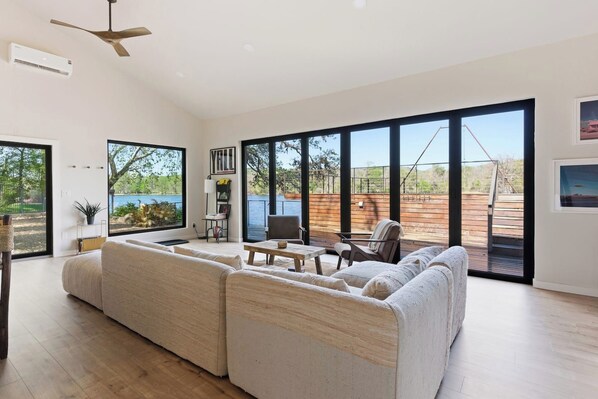 A spacious living room with a large sectional sofa, a dining table, and chairs. There are large windows offering a view of the outdoors.