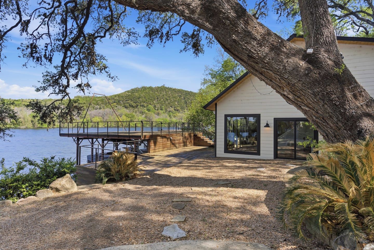 A lakeside cabin with a dock, a large tree in the foreground, and a view of the lake and hills in the background.