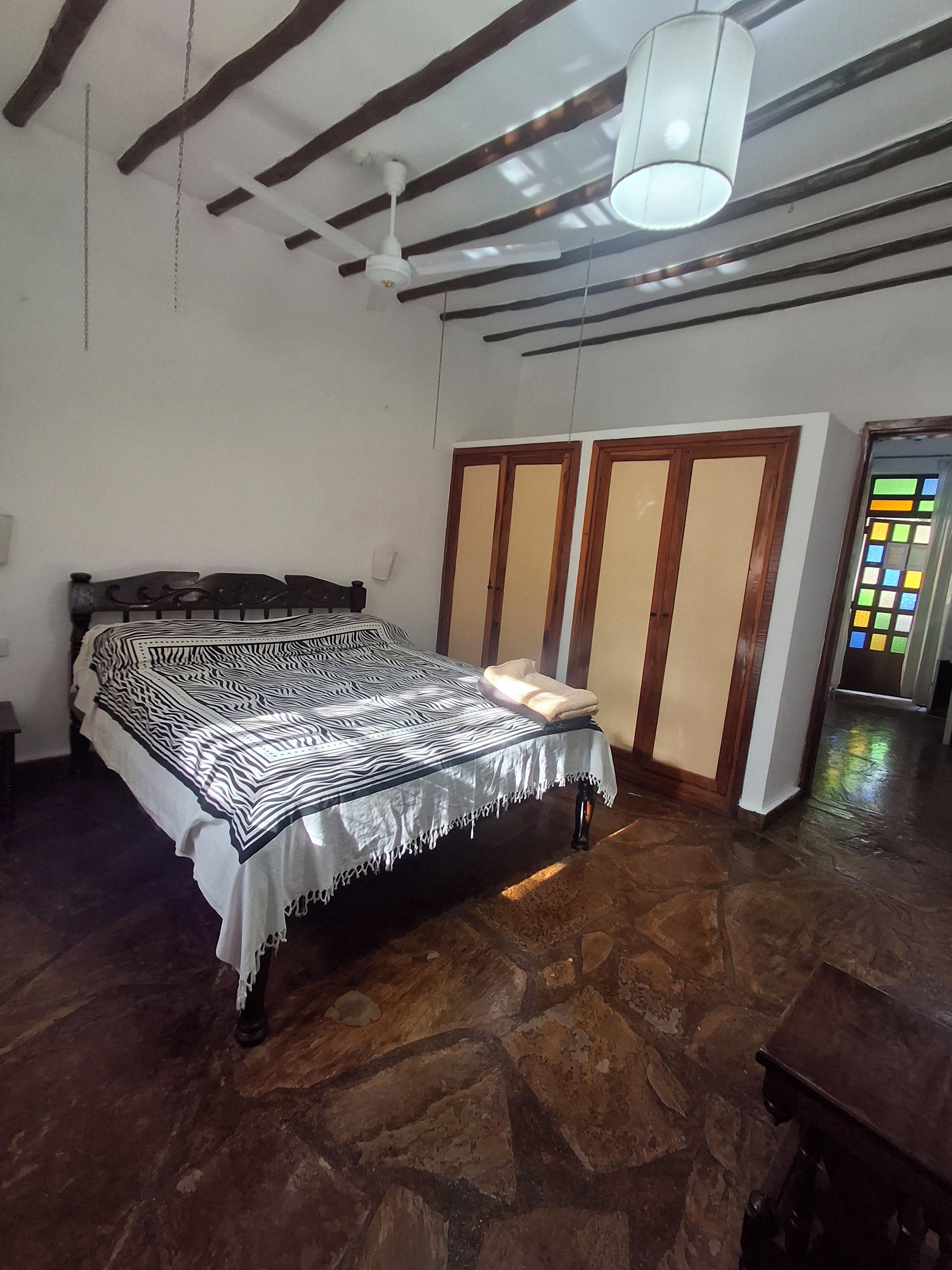 Een slaapkamer met een bed, een kledingkast, een plafondventilator en een stenen vloer.