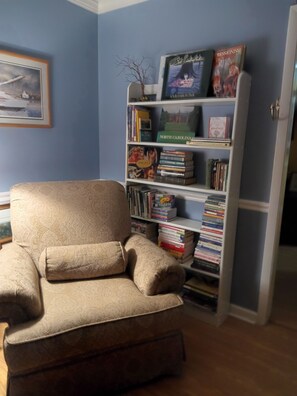 A room with a beige armchair, a bookshelf with various books and a framed picture, and a door leading to another room. - 3 Miles to Furniture Market • 8 Beds • Fast Wi‑Fi • Lake Views (High Point)