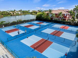Ein Tennisplatz im Freien mit mehreren Spielfeldern, umzäunt und mit einem Gewässer im Hintergrund.
