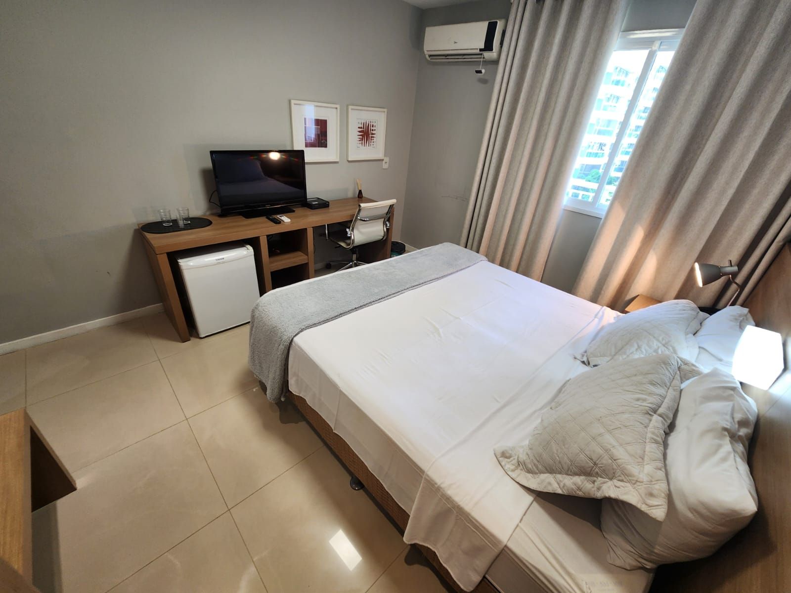 Et hotelværelse med seng, skrivebord, stol, lille køleskab og aircondition.