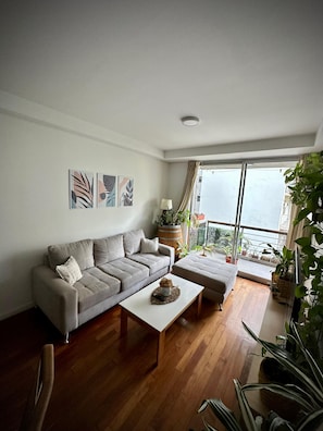 A living room with a sofa, a coffee table, and a view of the outdoors through large windows. - Confortable 3 Ambientes en Núñez, zona Segura, conectado y con Balcón (Buenos Aires)