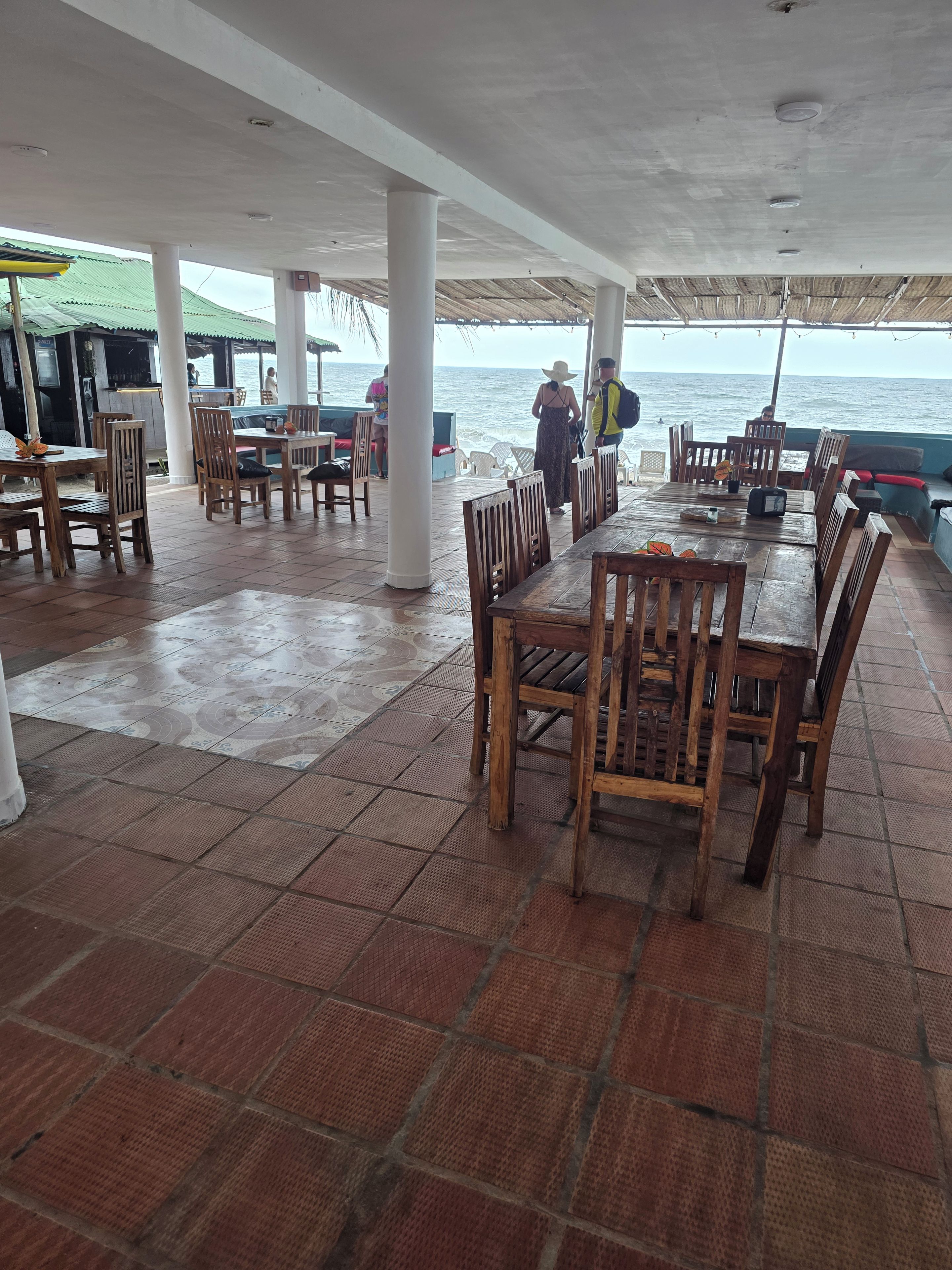 A covered outdoor dining area with wooden tables and chairs, a view of the sea, and a thatched roof structure.