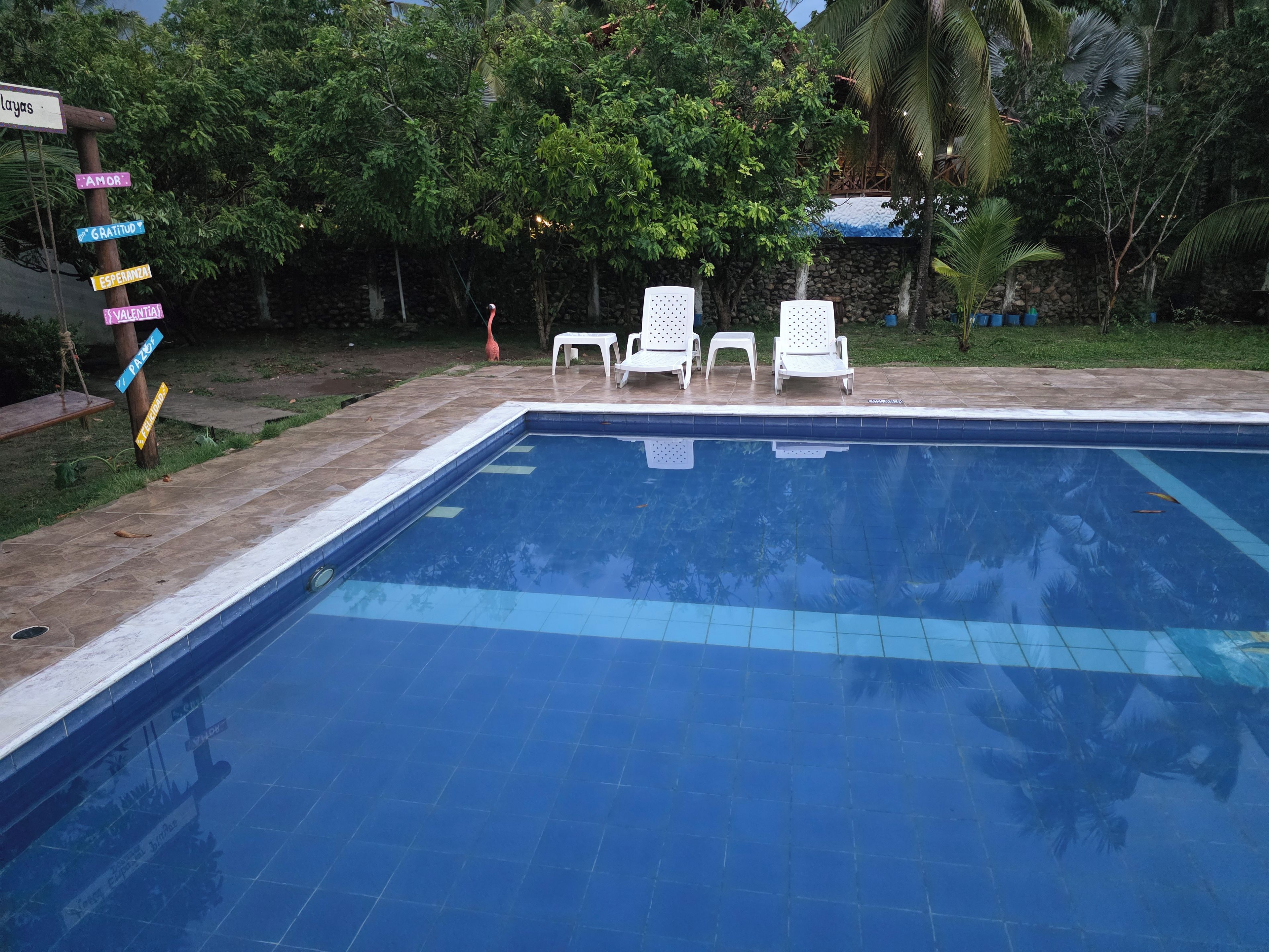 Piscina com borda azulejada, rodeada por espreguiçadeiras brancas, e um poste de sinalização com diversas placas indicativas.