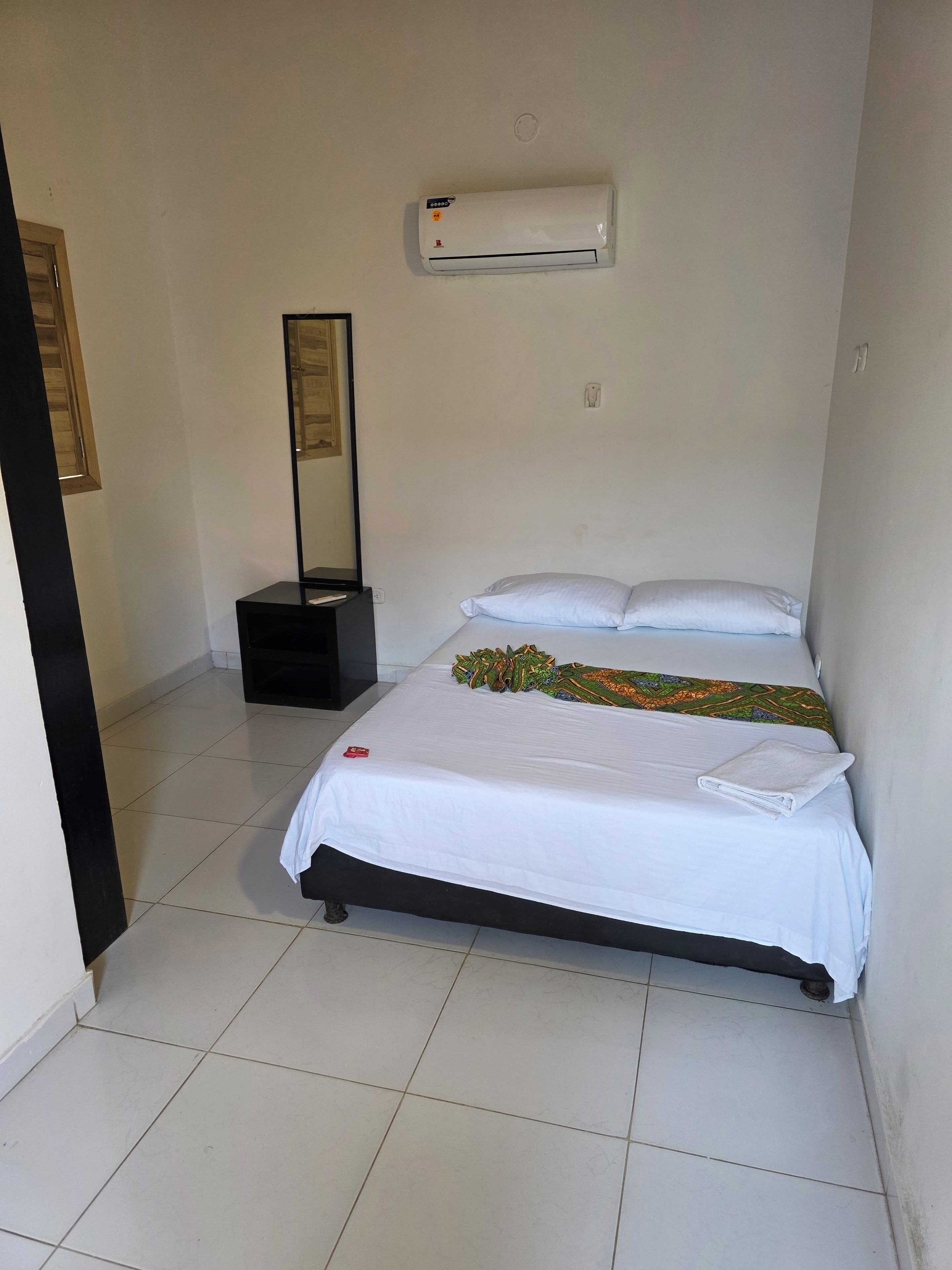 Habitación de hotel con una cama, aire acondicionado instalado en la pared, una mesita de noche con lámpara y una puerta que da a otra habitación.