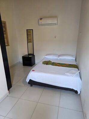 Habitació d'hotel amb llit, aire condicionat fixat a la paret, taula de nit amb làmpada i una porta que condueix a una altra habitació.
