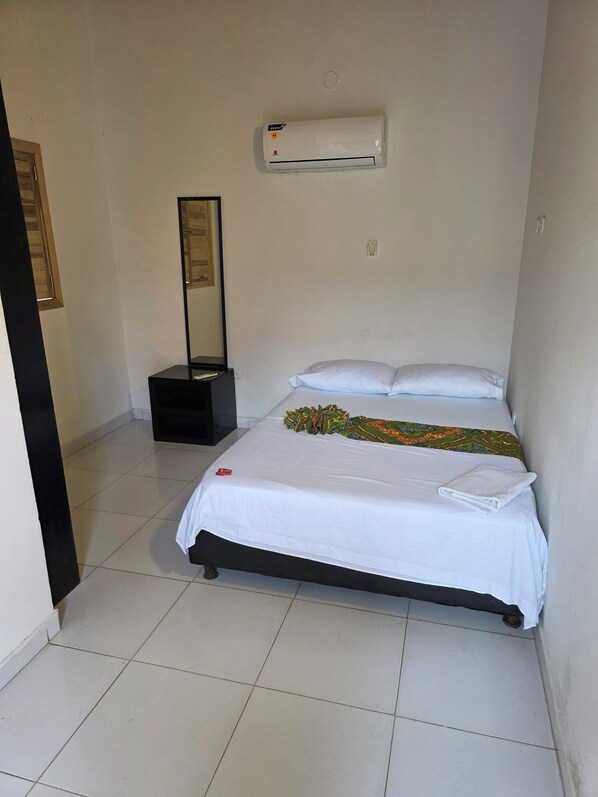 A hotel room with a bed, a wall-mounted air conditioner, a bedside table with a lamp, and a door leading to another room.