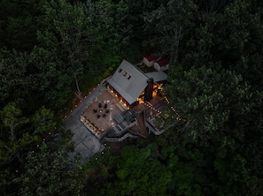 Eine Luftaufnahme einer Hütte, umgeben von dichtem Wald, mit einer Terrassenfläche, die Sitzgelegenheiten und eine Feuerstelle bietet.