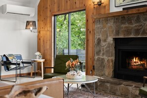 Ein gemütliches Wohnzimmer mit Kamin aus Stein, einem grünen Sessel, einem Glastisch mit Blumen und Blick ins Grüne.