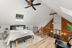 Een slaapkamer met een schuine kap, een bed met witte beddengoed, een bankje, een nachtkastje met een lamp, en uitzicht op groen door een raam.