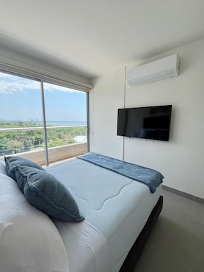 A bedroom with a bed, a TV mounted on the wall, and a large window offering a view of greenery and a bridge. - Cozy 1-bedroom Apartment - Condo Sea + Mountain Views (Santa Marta)
