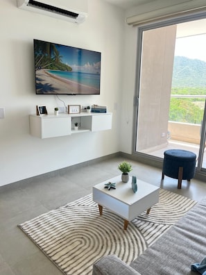 A modern living room with a flat-screen TV mounted on the wall, a white console table with decorative items, a blue ottoman, a striped rug, and a view of a mountainous landscape through a sliding glass door. - Cozy 1-bedroom Apartment - Condo Sea + Mountain Views (Santa Marta)
