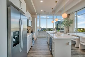 A modern kitchen with stainless steel appliances, a water dispenser, and a view of a cityscape through large windows. - Downtown/FreeParking/Rooftop Pool/ King Plush Beds (Atlanta)