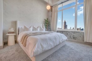 A modern bedroom with a large bed, a bedside table, a window with a city view, and a hanging light fixture. - Downtown/FreeParking/Rooftop Pool/ King Plush Beds (Atlanta)