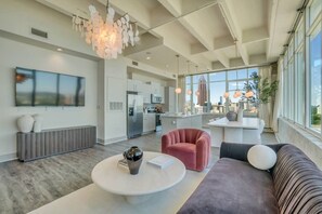 A modern living room with a large sofa, a coffee table, and a pink armchair. There is a kitchen area with a refrigerator and a dining table. The room has large windows and a view of the city. - Downtown/FreeParking/Rooftop Pool/ King Plush Beds (Atlanta)