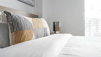 A neatly made bed with a striped pillow and a wooden headboard, a nightstand with a lamp, and a large window with a view of buildings.