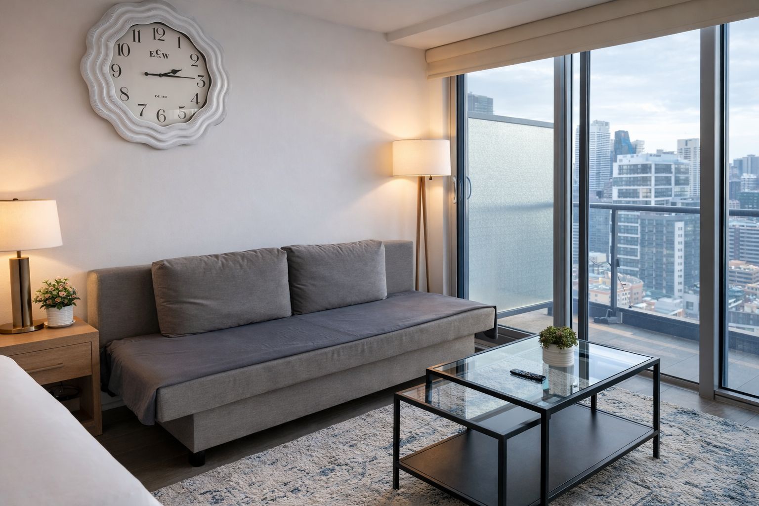 Et moderne hotelværelse med en grå sofa, et glasbord, et vægur og et stort vindue med udsigt over byen.