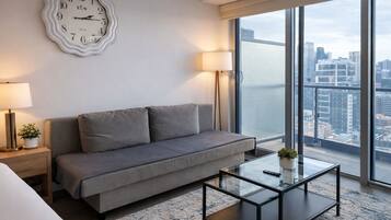 Et moderne hotelværelse med en grå sofa, et glasbord, et vægur og et stort vindue med udsigt over byen.
