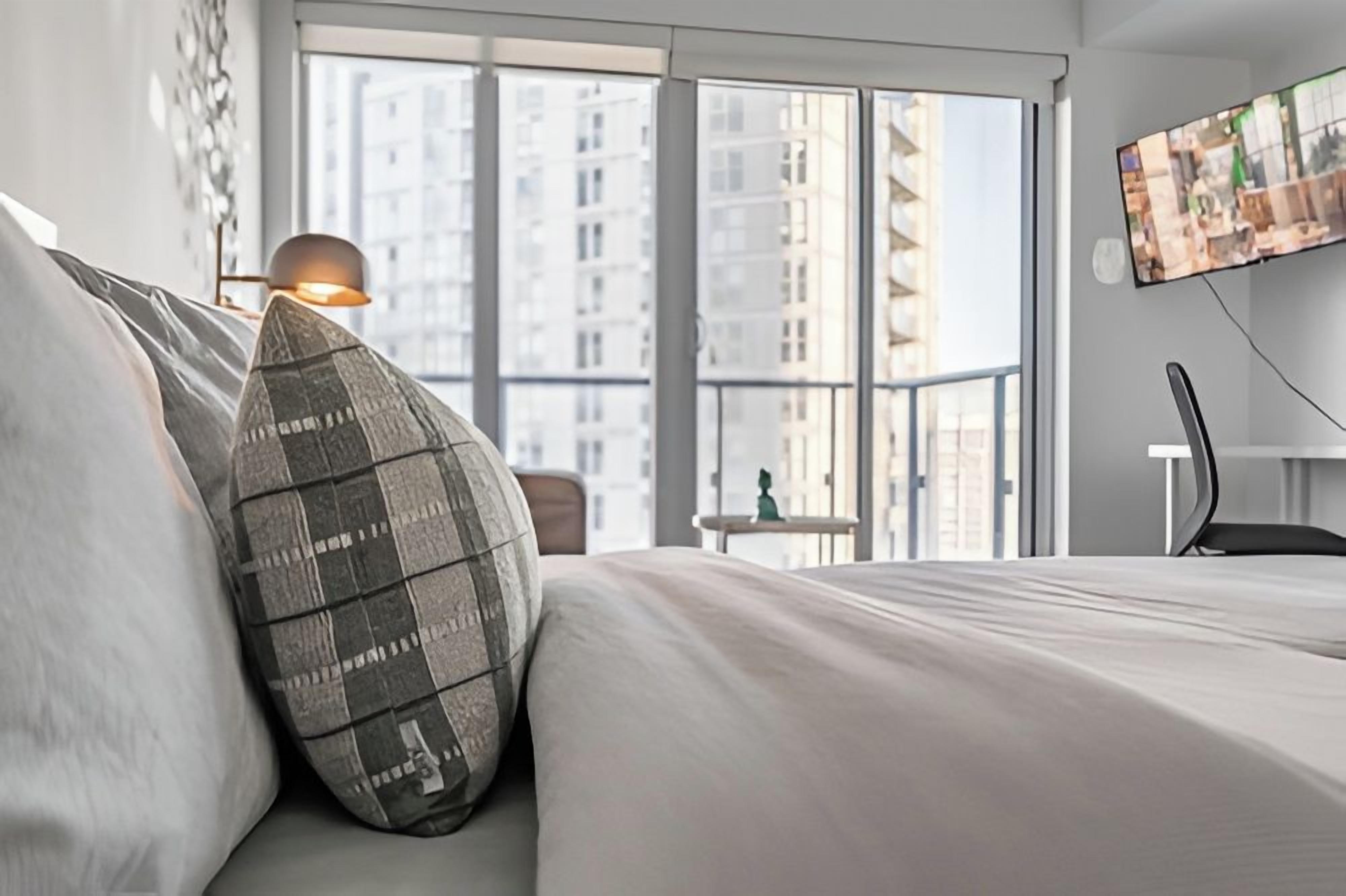 A modern hotel room with a large window, a bed with a pillow, a desk with a chair, and a TV mounted on the wall.