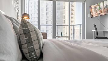 A modern hotel room with a large window, a bed with a pillow, a desk with a chair, and a TV mounted on the wall.