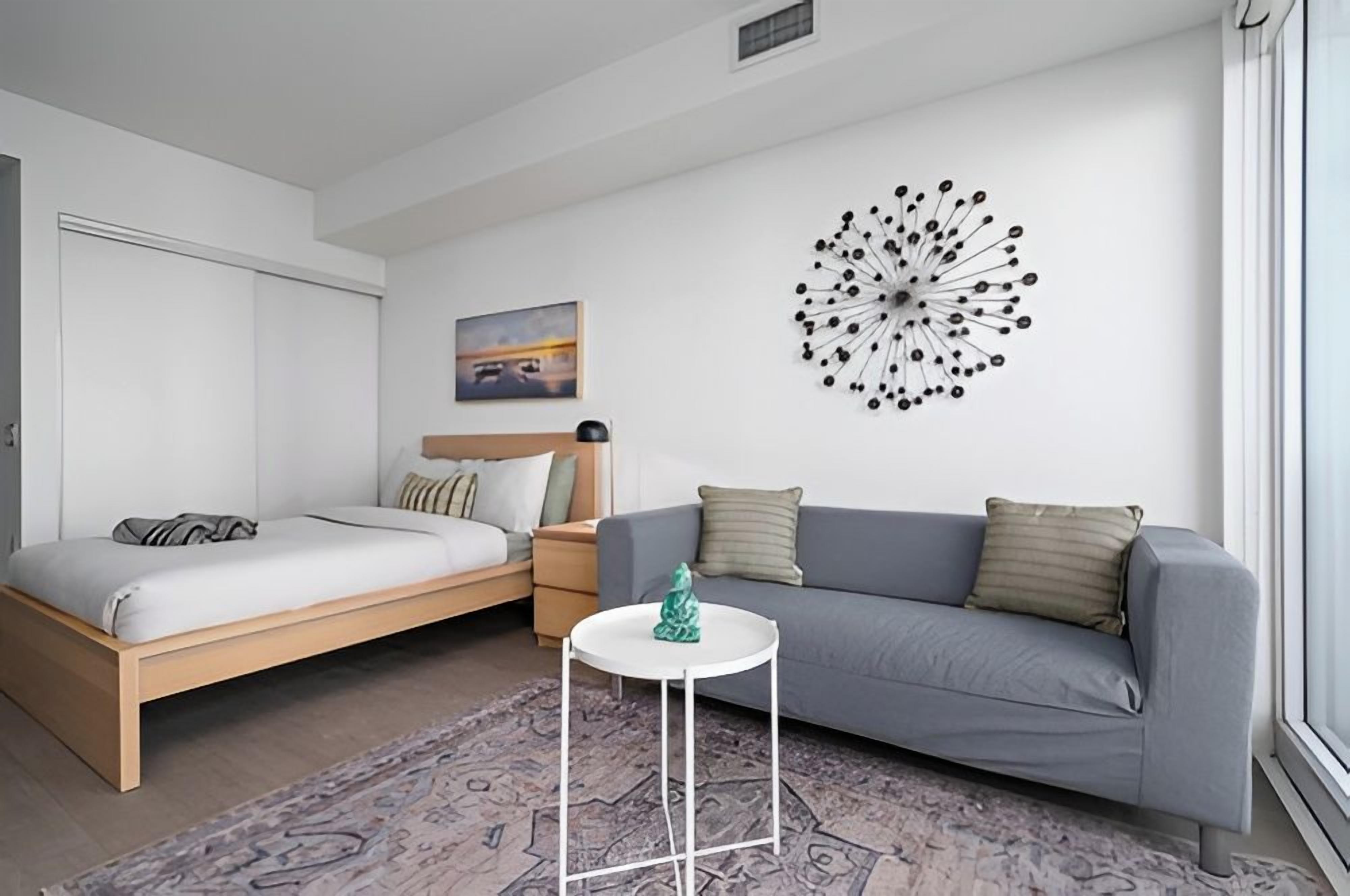 A modern bedroom with a bed, a grey sofa, a small white table, and a wall art piece.