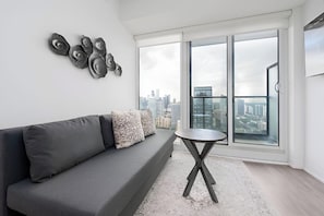 A modern living room with a gray sofa, patterned pillows, a round coffee table, and a city view through large windows. - The Boutique Suites - Jarvis Street (Toronto)