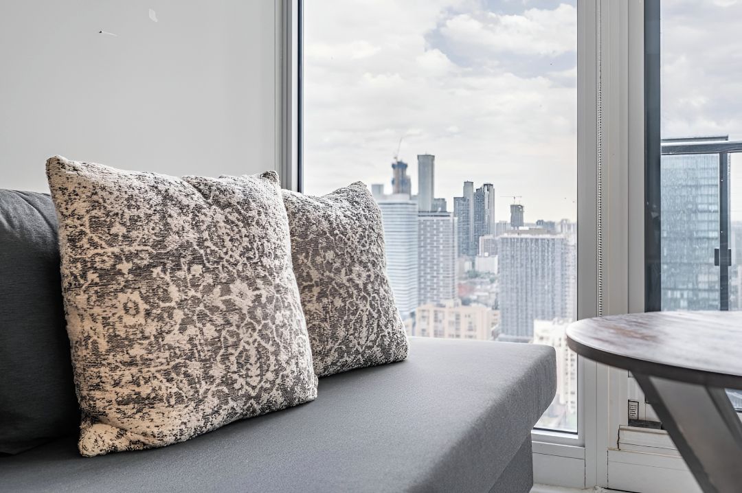 Una sala de estar moderna con un sofá y cojines, una mesa redonda y un ventanal con vistas a la ciudad.